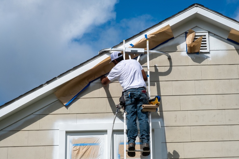 Worker on a ladder securing trim and soffit vent boards at the roofline of a house